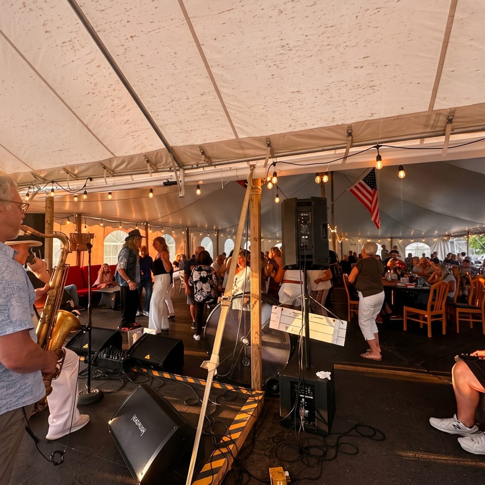 Crowded outdoor tent venue with a saxophonist performing, seated diners, string lights, speakers, and an American flag.