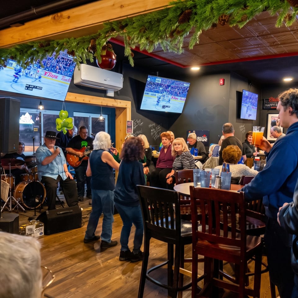 Crowded restaurant bar with a live band performing as patrons watch, dance, and enjoy drinks beneath holiday garlands and sports screens.