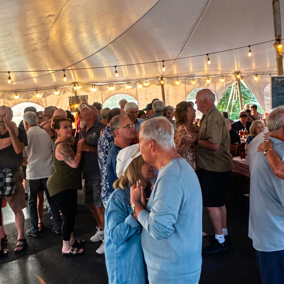 Guests dance beneath string lights inside a festive restaurant tent with picnic tables and a lively crowd.
